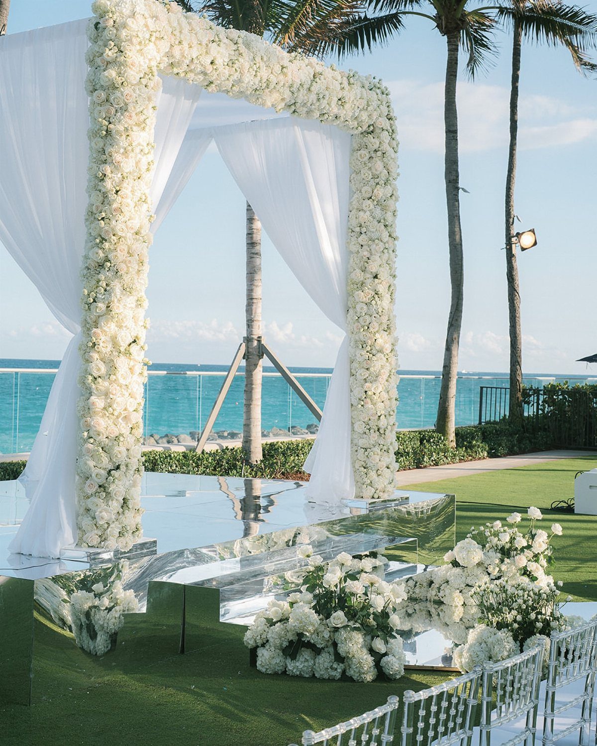 White floral wedding arch on a mirrored platform overlooking a turquoise ocean beach.