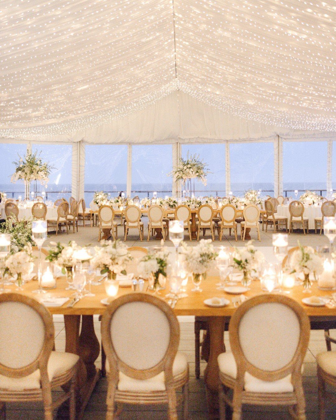 Wedding reception tables with white flowers under a lit tent overlooking the ocean.