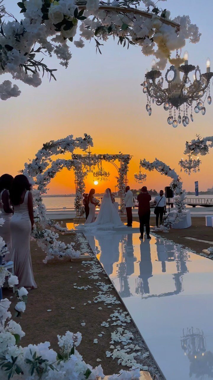 Wedding ceremony with white floral arches and reflective aisle overlooking water at sunset.