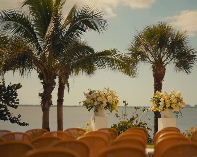 Waterfront wedding ceremony setup with orange chairs, white flowers, and palm trees.
