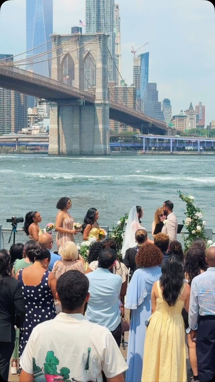 Wedding ceremony on a waterfront pier with a large bridge and city skyline background.