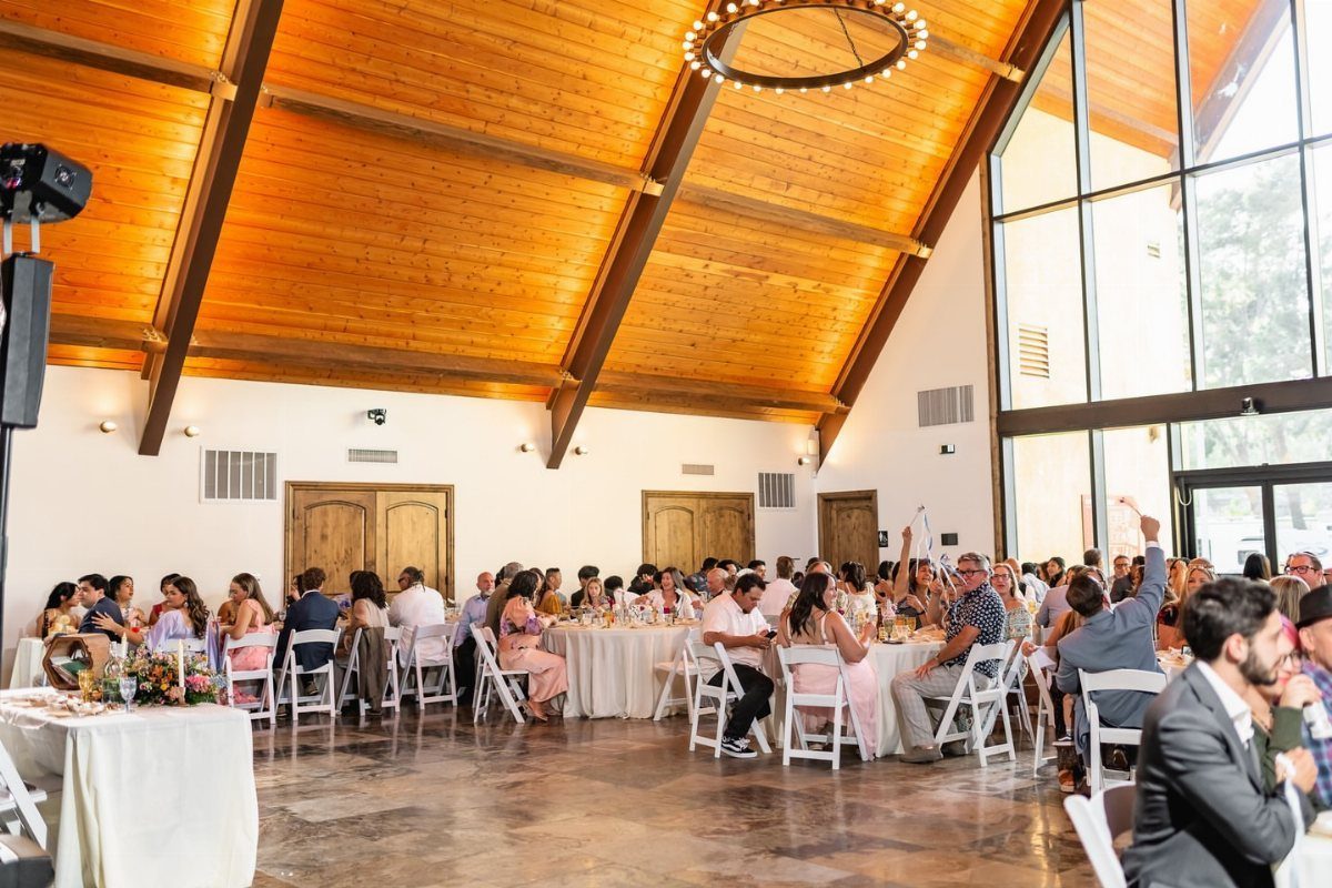 Wedding reception guests seated at round tables in a hall with wooden ceilings.