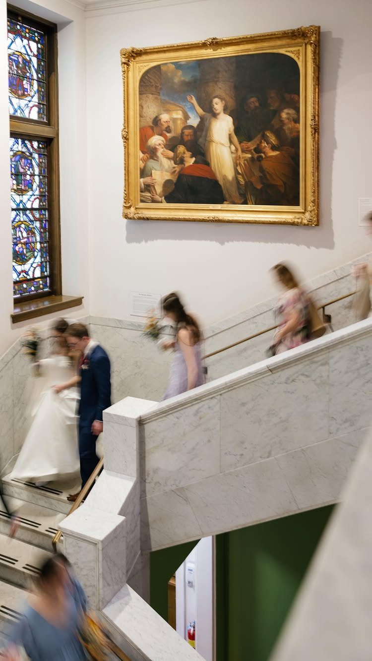 Bride and groom walking down a marble staircase beneath a large classical painting.