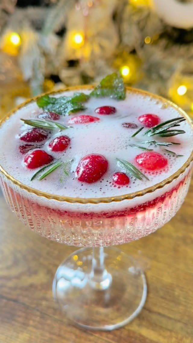Pink cocktail with cranberries and rosemary in a gold-rimmed glass with festive lights.