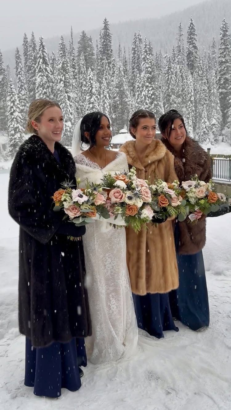 Bride and bridesmaids in fur coats holding bouquets in a snowy pine forest.