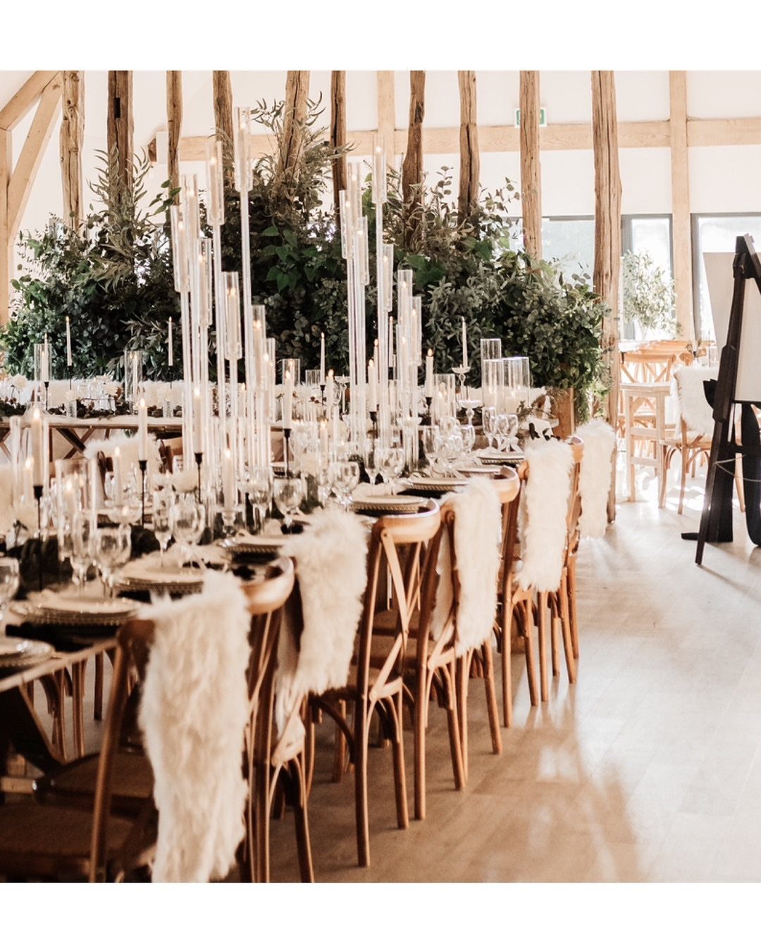Rustic barn wedding table with tall white candles and faux-fur draped wooden chairs.