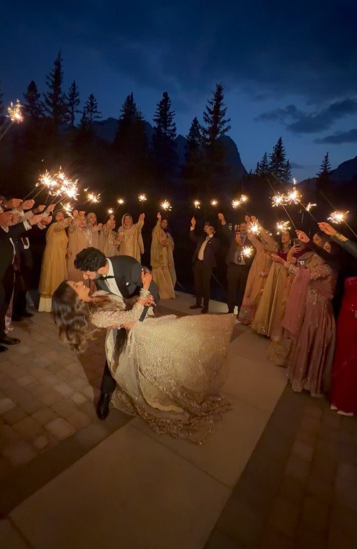 Groom dipping bride for a kiss while guests hold sparklers in a winter forest