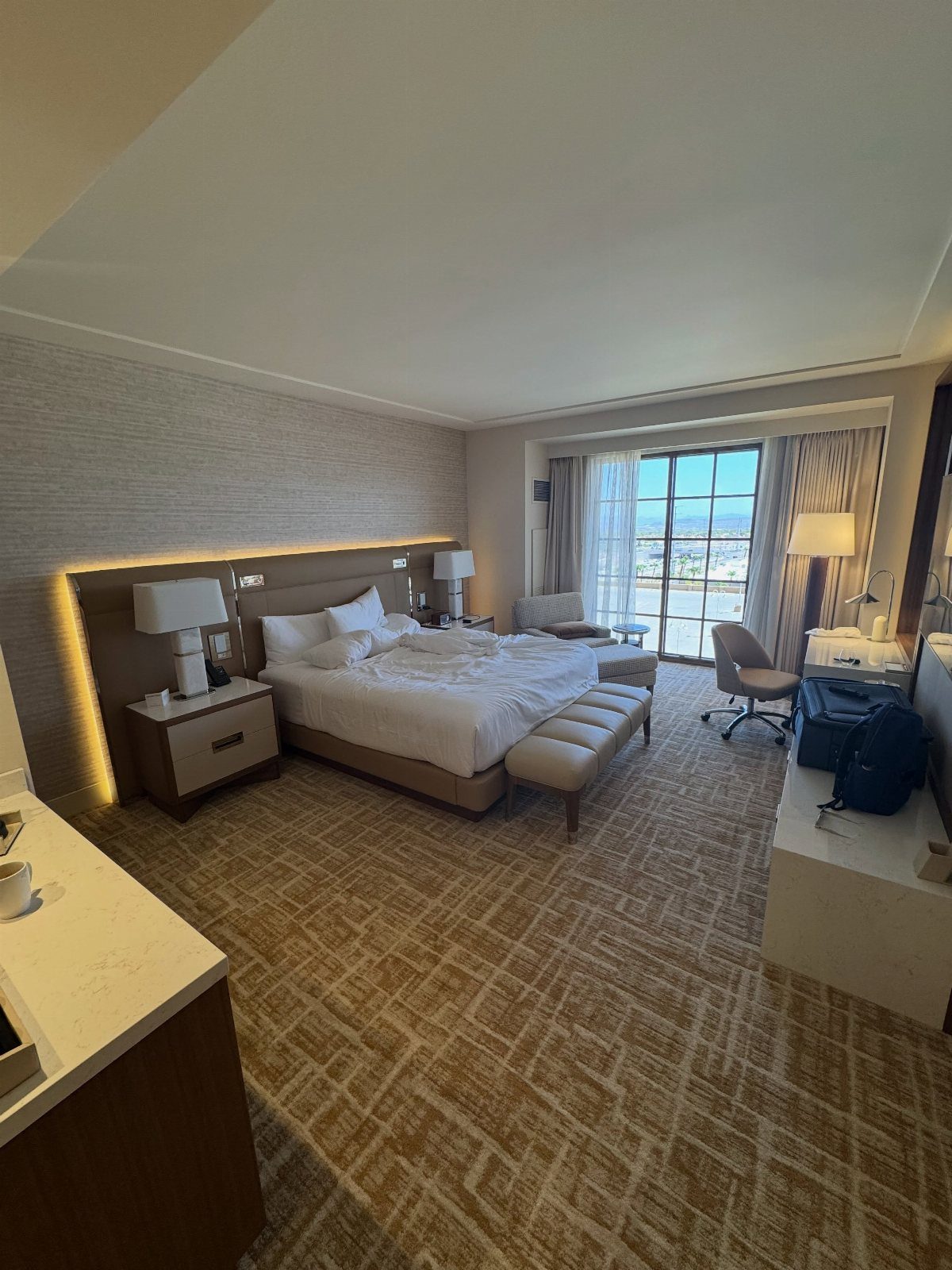 Modern hotel suite with a large white bed and city view window.